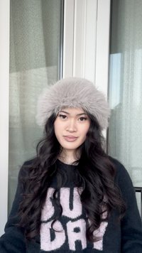 Noor naine, kellel on pikad lokkis juuksed, kannab tuhmunud halli karvaseid mütsi ja musta kampsunit, millel on suured valged plokk tähed, seisab toas akna lähedal.