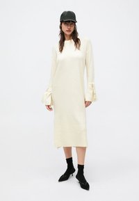 Kremowa sukienka z prążkowanego dzianiny z długimi rękawami, ozdobiona kokardkami przy mankietach. W zestawie z czarną czapką i spiczastymi butami.