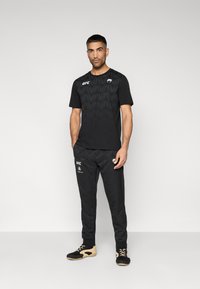 Μαύρο αθλητικό t-shirt με γεωμετρικό μοτίβο, λογότυπο UFC στο στήθος· συνδυασμένο με μαύρο παντελόνι, ελαστικές μανσέτες και λογότυπο και στα δύο κομμάτια.