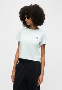 Γυναίκα με πλούσια σγουρά μαλλιά, φορώντας στρογγυλά μαύρα γυαλιά ηλίου, ένα ανοιχτό μπλε cropped T-shirt της North Face και μαύρο παντελόνι, μπροστά σε απλό φόντο.