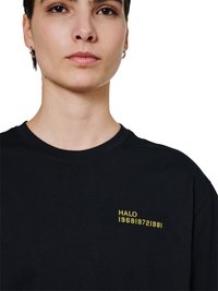 Młoda osoba nosząca czarną koszulkę z okrągłym dekoltem z małym żółtym napisem "HALO 1968|972|981" na lewej piersi, na białym tle.