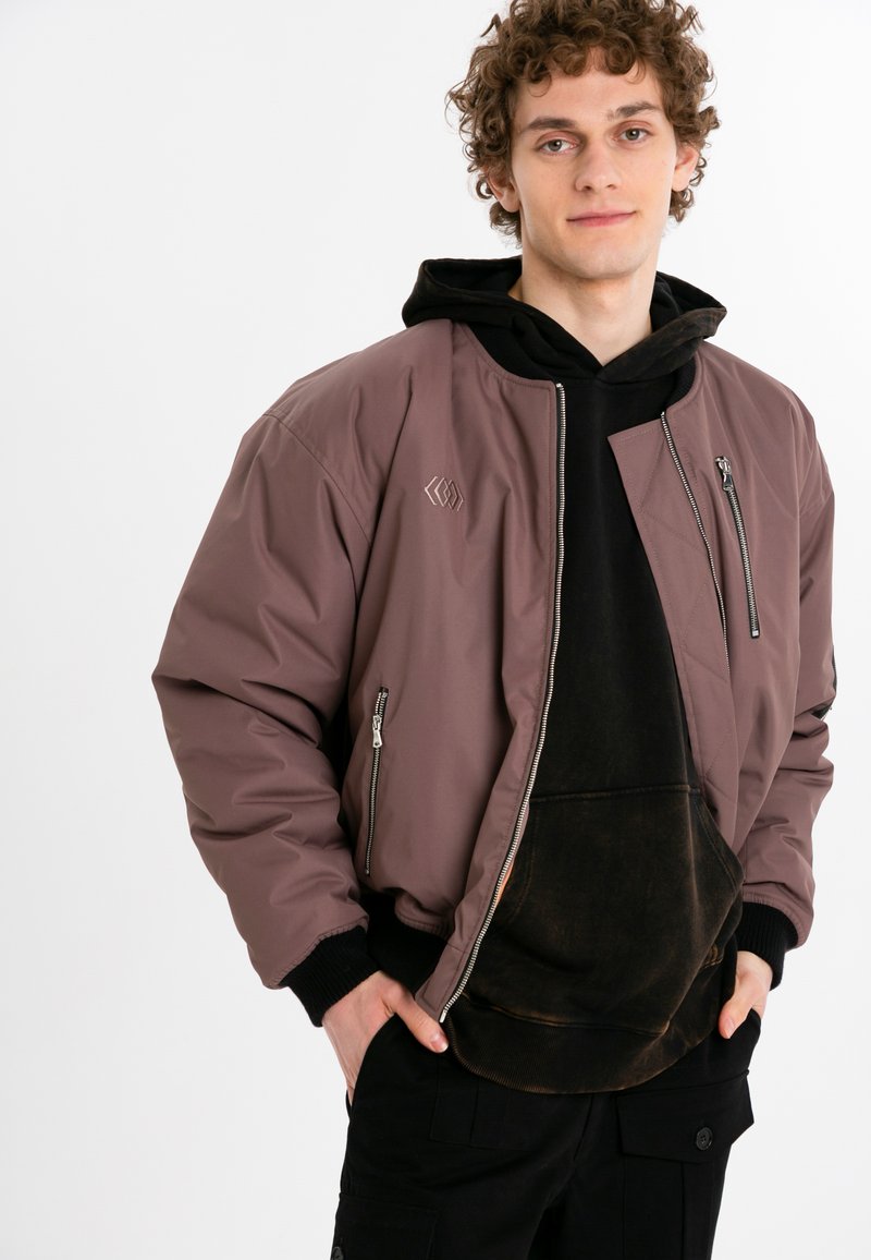 Brun bomberjacka med dragkedja framtill, ribbad mudd och logodetalj. Lager på en svart hoodie med en urblekt textur.