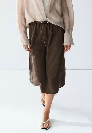 Persona che indossa una gonna ampia marrone scuro con coulisse, una camicia beige trasparente e sandali infradito bianchi mentre cammina su un pavimento bianco.