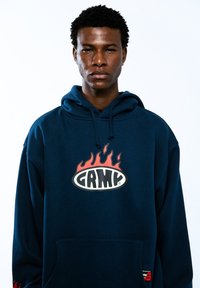 Sudadera con capucha de color azul marino hecha de tejido suave, con un logo central que presenta llamas y la palabra "GRMY". Incluye un bolsillo tipo canguro y puños de canalé.