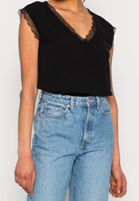 Haut noir à manches courtes en V avec bord en dentelle, associé à un jean en denim bleu clair taille haute avec cinq poches et une fermeture à bouton.