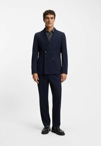 Marinblå dubbelknäppt kavaj med slagiga kragar och sidosfickor, kombinerad med matchande byxor och svarta loafers. Prickig skjorta under.