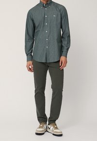 Camisa de botones verde a rayas con cuello, combinada con pantalones oliva y zapatillas beige con acentos negros.