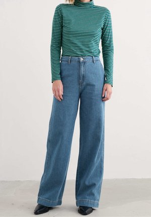 Personne portant un col roulé rayé vert et noir à manches longues, un jean bleu taille haute à jambes larges, et des chaussures noires pointues, debout sur un sol clair.