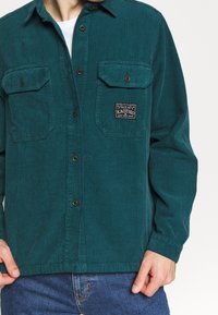 Teal kordflorskjorta med knapp-up framsida, två bröstfickor och sydd etikett, med ribbad textur och lång ärm.