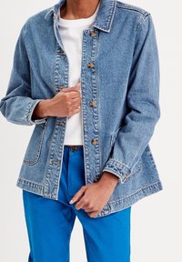 Helles blaues Denim-Jackett mit klassischem Kragen, Knopfverschluss, zwei vorderen Taschen und leicht übergroßem Schnitt. Über einem weißen Shirt und blauen Hosen getragen.