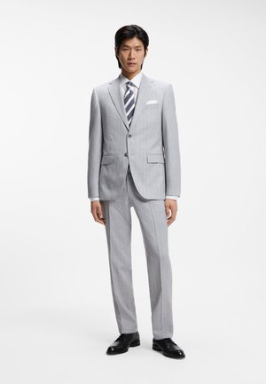 Abito a righe grigio con design aderente, pantaloni coordinati, camicia bianca e cravatta a righe. Scarpe eleganti nere completano il look.