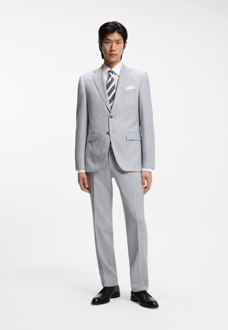 Grauer gestreifter Anzug mit figurbetontem Schnitt, passende Hose, weißes Hemd und gestreifte Krawatte. Schwarze Anzugschuhe vervollständigen das Outfit.
