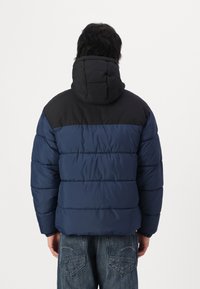 Puffer jacket in navy blue with a black top panel. Features horizontal stitching, a hood, and a soft, insulated texture.