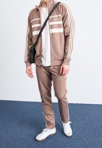 Chándal deportivo marrón con rayas blancas, chaqueta con cremallera, pantalones a juego y zapatillas blancas. Bolsa de hombro negra con detalles verdes.