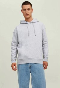 Jovem com um hoodie cinza claro com cordões e bolso frontal, combinado com jeans azuis claros, pousando sobre um fundo simples.