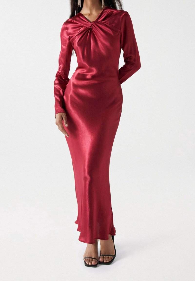 Czerwona satynowa sukienka z długim rękawem, z plisowanym dekoltem i dopasowanym krojem, z rozszerzanym dołem i w zestawie z czarnymi sandałami na szpilkach.