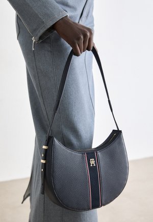 Ręka trzymająca czarną, teksturowaną skórzaną torbę na ramię z złotym logo i detalem z czerwono-paskowanej tkaniny, ubrana w szary płaszcz z rękawami na suwak.