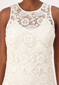 Mouwloos kanten topje in ivoorkleur met bloemmotieven, featuring een textuurontwerp en een ronde halslijn. Fijne haakdetails door het hele jaartje.
