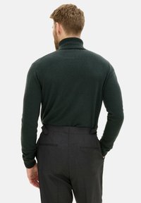 Dunkelgrüner, dick gestrickter Pullover mit hohem Kragen. Getragen mit dunkelgrauen, maßgeschneiderten Hosen mit seitlichen Taschen und hinteren Akzenten.