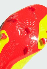 Suela de botas de fútbol roja y amarilla con tachones triangulares elevados, superficie texturada y "RATOR" grabado en un lado. Bordes curvados.