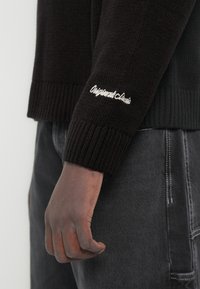 Σκούρο δέρμα χεριού ακουμπά δίπλα σε μανίκι μαύρου πουλόβερ πλεκτού με λευκή κεντητή επιγραφή «Originals Shorts», σε συνδυασμό με σκούρο γκρι τζιν.