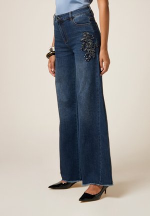 Jeans in denim scuro a vita alta con gambe ampie e dettagli floreali ricamati sulla coscia sinistra. Presentano un orlo sfrangiato e un classico design a cinque tasche.