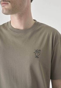 Olive green cotton t-shirt with a round neckline and a small black bird graphic on the left chest. Smooth texture, short sleeves.