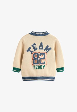 Kremowy sweter z dzianiny z niebieskimi i zielonymi ściągaczami na mankietach i kołnierzu, z napisem "TEAM 82 TEDDY" w kolorach niebieskim, czerwonym i zielonym na plecach.