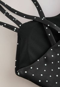 Masque facial noir à pois blancs, avec plusieurs fines lanières pour un maintien sûr sur un fond clair.