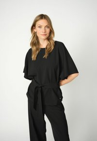 Top nero con maniche larghe e vita annodata, realizzato in un tessuto liscio. Abbinato a pantaloni neri, con un sottile dettaglio del logo.