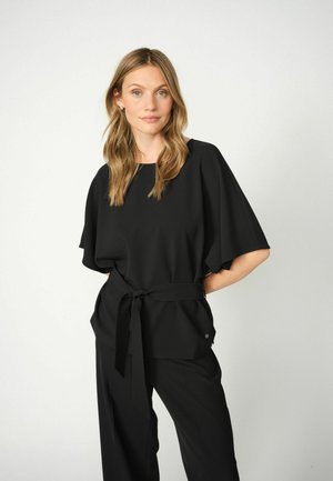 Top nero con maniche larghe e vita annodata, realizzato in un tessuto liscio. Abbinato a pantaloni neri, con un sottile dettaglio del logo.