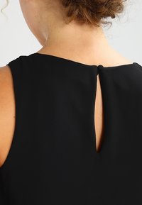 Persona de cabello rizado que lleva una blusa sin mangas de color negro con un pequeño abertura en la parte de atrás, cerrada con un botón cubierto de tela.