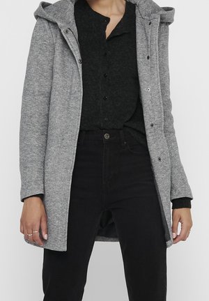 Person trägt einen grauen Kapuzenmantel über einem schwarzen, geknöpften Pullover und schwarzen, hoch taillierten Hosen, steht vor einem schlichten Hintergrund.