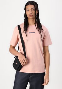 Lyserød T-shirt med "element" logo i marineblå, sammen med mørke jeans og en lille sort crossover taske. Afslappet pasform, kortærmet.