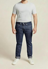 Homme portant un jean bleu foncé ajusté avec les revers roulés, un t-shirt gris clair uni et des baskets blanches, debout devant un fond neutre.
