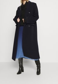 Dubbelknäppt marinblå kappa med breda slag, texturerad svart stickad tröja, plisserad blå kjol och svarta knähöga stövlar.