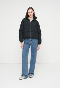 Μαύρο φουσκωτό μπουφάν με φερμουάρ μπροστά και ψηλό γιακά, με μοτίβο καπιτονέ. Συνδυασμένο με μπλε παντελόνι καμπάνα και ασημένια παπούτσια.