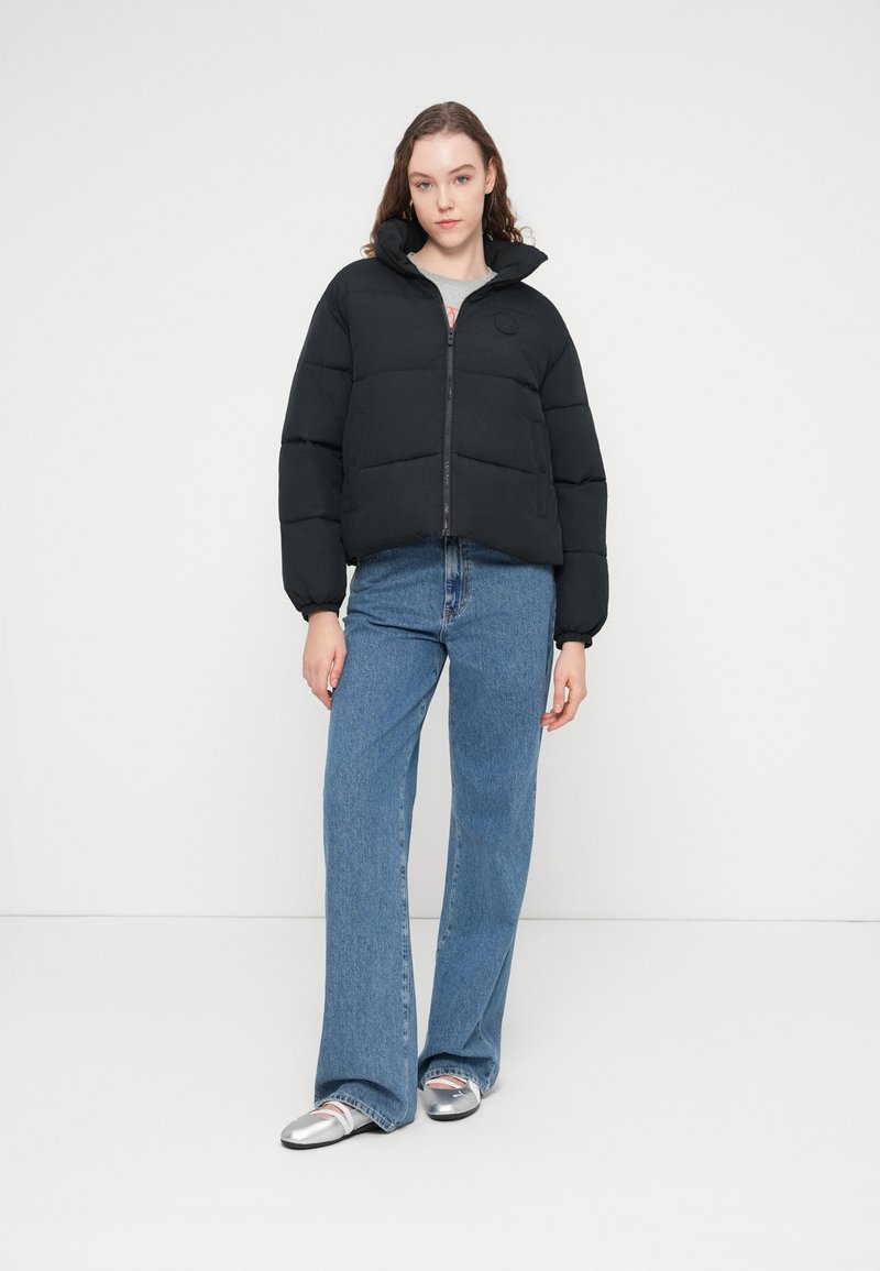 Sort dunjakke med lynlås foran og høj krave, med et quiltet design. Kombineret med blå flared jeans og sølvfarvede sko.