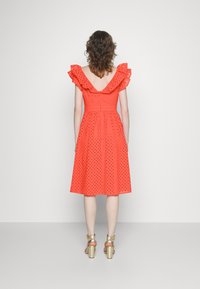 Vestido vermelho até ao joelho com mangas franzidas, costas abertas e padrão perfurado, combinado com sandálias de salto alto douradas.