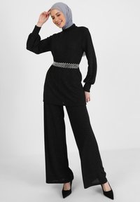 Tunika nera e luccicante con collo alto e maniche lunghe, abbinata a pantaloni a gamba larga. Accentuata da una cintura con gioielli e scarpe con tacchi alti.