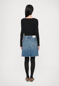 Femme aux longs cheveux noirs de dos, portant un haut noir à manches longues, une jupe en jean bleue, des collants noirs et des chaussons imprimés léopard.