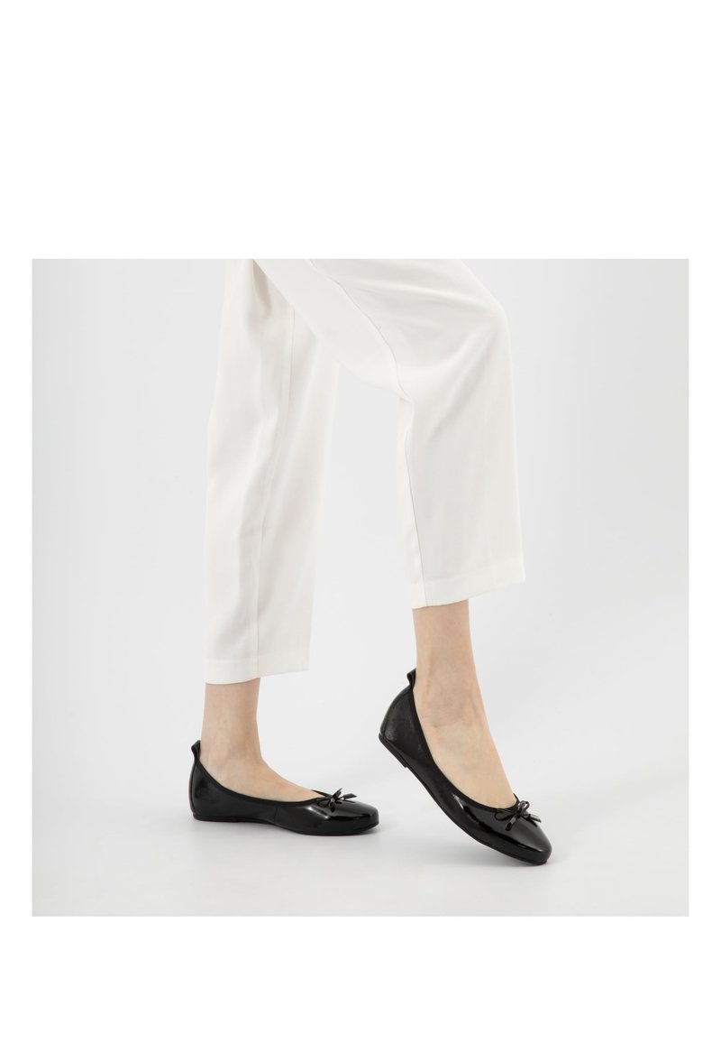 Kobieta ubrana w białe spodnie i czarne baleriny z małymi kokardkami, stojąca na jasnoszarym tle.