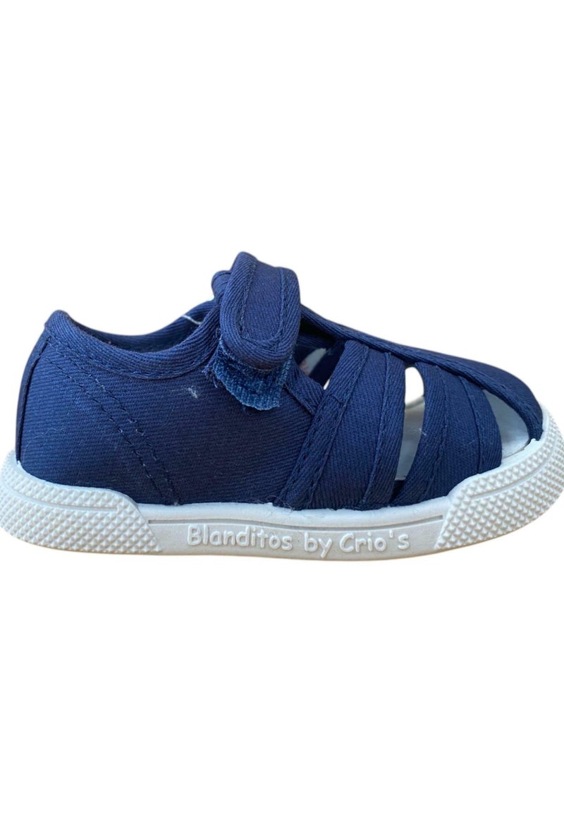 Zapato infantil de lona azul marino con cinta de velcro, punta abierta, diseño entrecruzado, suela de goma blanca y detalles de agarre texturizados.