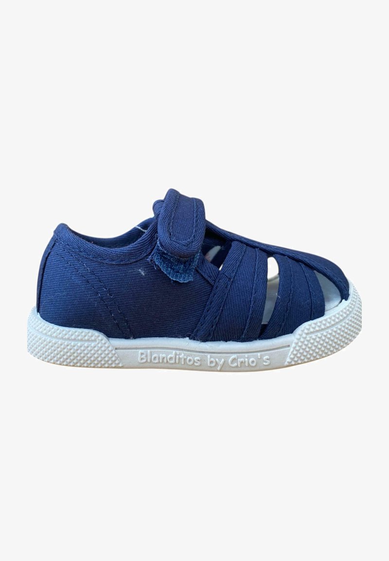 Zapato infantil de lona azul marino con cinta de velcro, punta abierta, diseño entrecruzado, suela de goma blanca y detalles de agarre texturizados.
