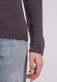 Main détendue le long du corps, portant un pull en maille violet foncé et un jean en denim bleu clair sur un fond uni.