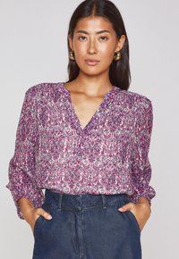 Purple and white patterned blouse with a V-neck, long sleeves, and lightweight fabric, paired with dark denim pants.