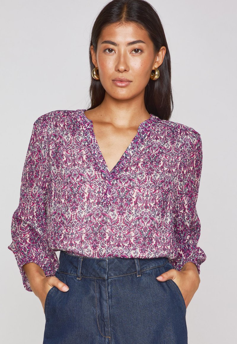 Purple and white patterned blouse with a V-neck, long sleeves, and lightweight fabric, paired with dark denim pants.