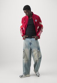 Giacca rossa con zip e strisce bianche, canotta nera, jeans cargo blu chiaro con tasche e sneakers con fantasia. Sfondo semplice.
