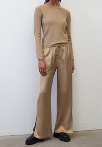 Lichtbeige ribbeltrui met lange mouwen, gecombineerd met losse, glanzende, wijd uitlopende tan satijnen broeken, met een trekkoord in de taille en zijsplitten. Zwarte laarzen met een dikke zool.