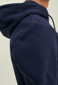 Felpa blu navy con una consistenza morbida, dotata di cappuccio e cordoncino. Inquadratura ravvicinata dell'area della spalla, evidenziando i dettagli del tessuto e delle cuciture.